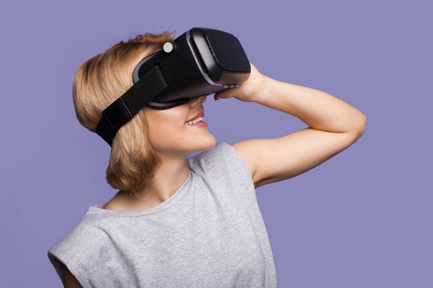 Foto vista lateral de una mujer caucásica con el pelo rubio sonriendo usando auriculares de realidad virtual sobre un fondo púrpura - Foto, Imagen