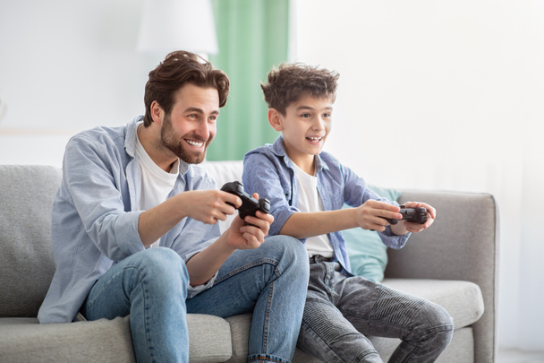 Chłopcy to chłopcy. Radosny ojciec i nastoletni syn rywalizują ze sobą w grach wideo online, używając joysticków - Zdjęcie, obraz