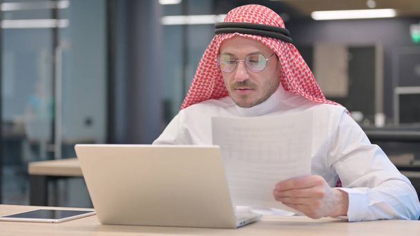 Hombre árabe de mediana edad con documentos de lectura para computadora portátil - Foto, imagen