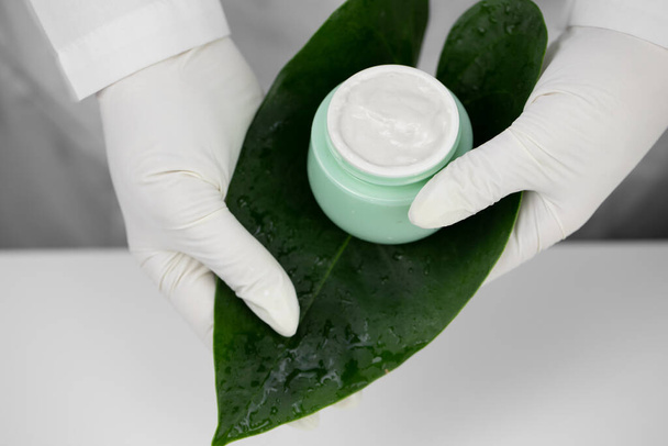 Крупным планом руки в медицинских перчатках держать банку крема на зеленом листе, белый фон - Фото, изображение