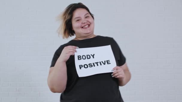 Concepto de positividad corporal - una mujer sonriente con sobrepeso bailando y sostiene un cartel con la inscripción CUERPO POSITIVO - Imágenes, Vídeo