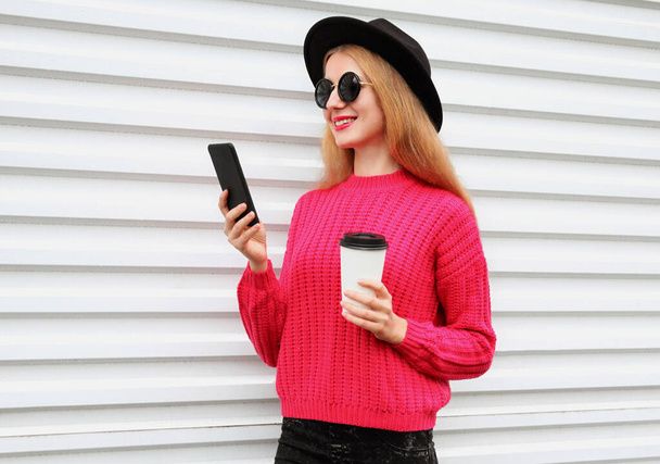 Portrait of stylish young woman with smartphone and cup of coffee on a city street wearing a colorful pink knitted sweater and black round hat on a white background - Photo, Image