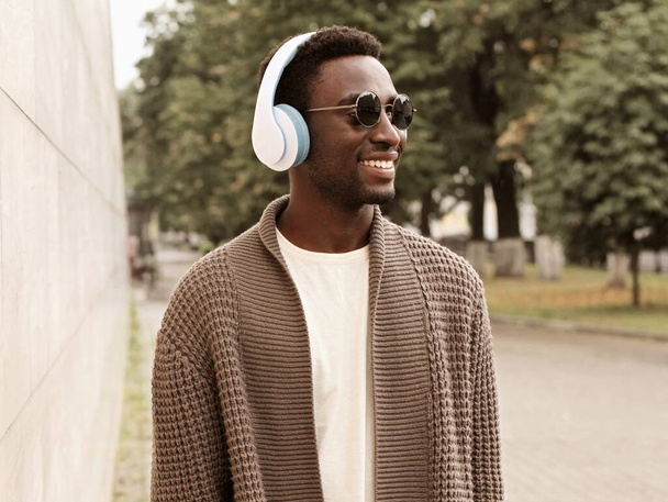 Portrait of happy smiling young african man in headphones listening to music wearing a brown knitted cardigan in a city - Foto, Imagem