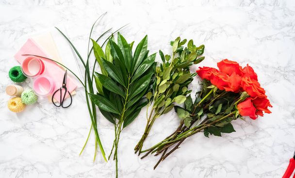 Flat lay. Arranging red roses and green leaves into a bouquet on a marble background. - Foto, Imagen