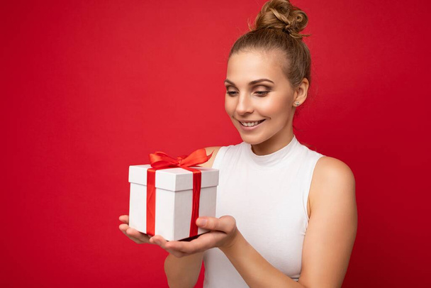 Photo shot of beautiful positive smiling young blonde woman isolated over red background wall wearing white top holding gift box and looking at present - Photo, Image