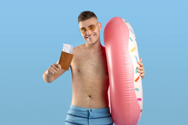 Handsome young guy with inflatable ring showing passport with tickets, going on tropical vacation over blue background - Valokuva, kuva