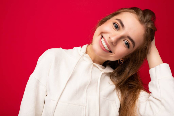 Jonge glimlachende mooie donkerblonde vrouw met oprechte emoties geïsoleerd op de achtergrond muur met kopieerruimte dragen casual witte hoodie. Positief concept - Foto, afbeelding