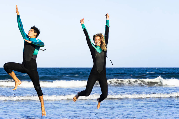 Kaksi nuorta paria hyppää sinisellä merellä. Ulkona kuva upea onnellisia ystäviä märkäpuku vitsi. Keskity tyttö kasvot. - Valokuva, kuva