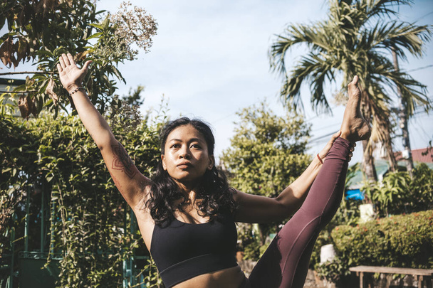 Egy fitt, fókuszált és cserzett ázsiai nő, aki az egyik lábán egyensúlyozik, miközben a szabadban jógázik egy napos reggelen.. - Fotó, kép
