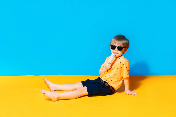 Zomer en vakantie in 2021. Gemengde race jongen in een zonnebril likt een ijsje. De blauwe achtergrond toont de lucht, en de gele achtergrond toont het zand. - Foto, afbeelding