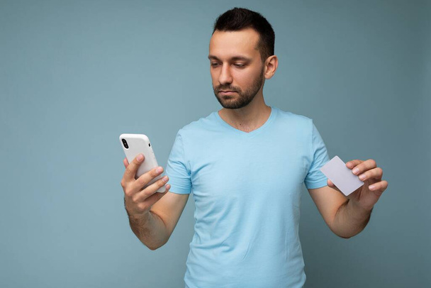 Photo of handsome young unshaven brunet man with beard wearing everyday blue t-shirt isolated over blue background holding and using mobile phone and credit card making payment looking to the side - 写真・画像