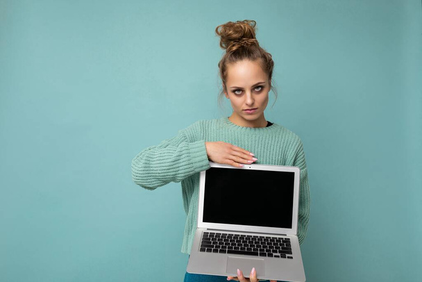 Komoly gyönyörű, sötét szőke fiatal nő összegyűjtött göndör haj nézi a kamerát tartó számítógép laptop üres monitor képernyő modellt, és másolja a helyet viselő kék hosszú ujjú elszigetelt - Fotó, kép
