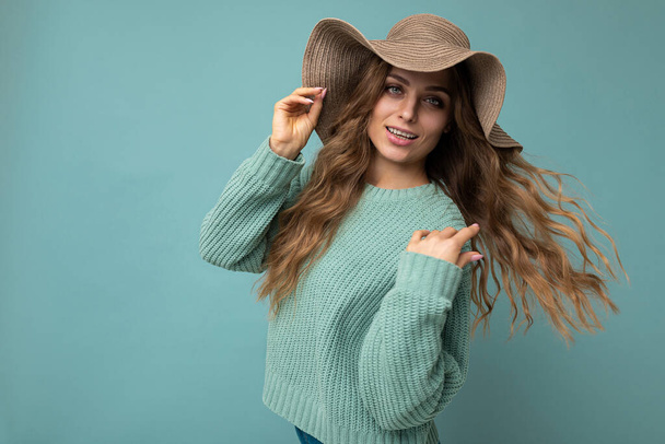 Portret młodej pięknej, uśmiechniętej hipsterki w modnym stroju i kapeluszu. Seksowna, beztroska kobieta pozująca w studio obok niebieskiej ściany z wolną przestrzenią. Pozytywny model z naturalnym makijażem - Zdjęcie, obraz