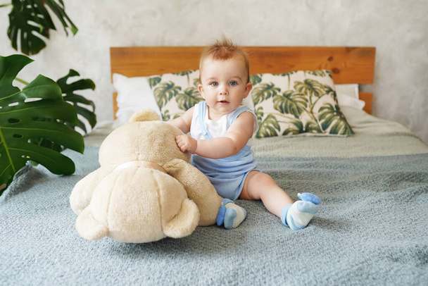 Adorable bébé garçon dans la chambre blanche ensoleillée tenant jouet ours en peluche avec plante verte monstera. Nouveau-né relaxant au lit. Textile et literie pour enfants. Matin de famille à la maison - Photo, image