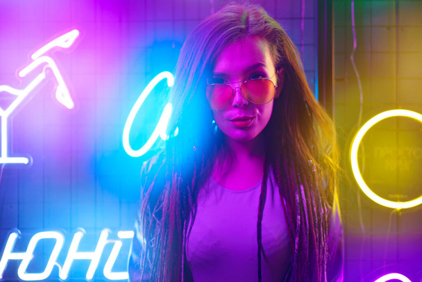 Divatportré egy fiatal nőről napszemüvegben, neon táblák mellett pózolva az éjszakai klubban. - Fotó, kép