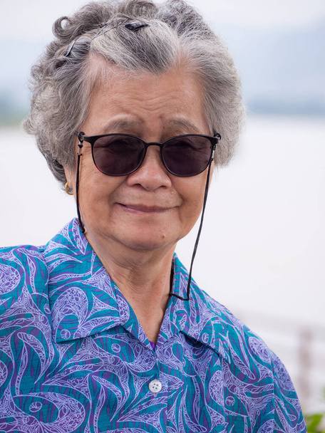 Portret van een bejaarde vrouw met kort wit haar die een zonnebril draagt en glimlacht aan de oever van de rivier. Begrip "ouderen" en "reizen" - Foto, afbeelding