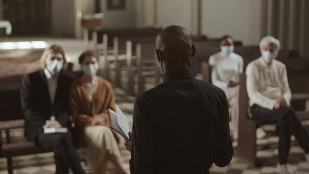 Vista posteriore del pastore afro-americano che indossa vestiti neri e maschera medica in piedi nella chiesa luterana con la Sacra Bibbia in mano e parla con i parrocchiani che indossano maschere - Filmati, video