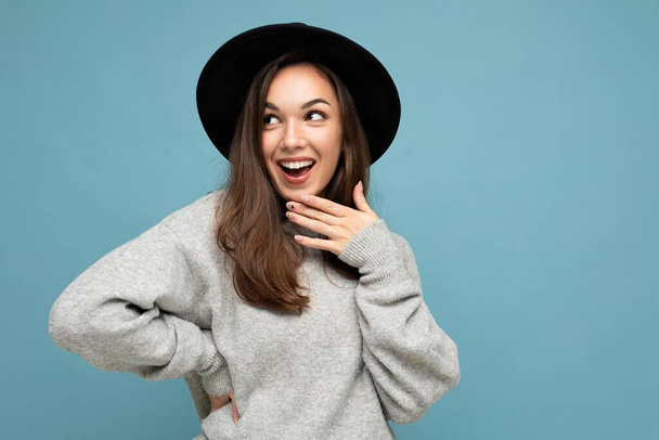 Portrait of young positive happy surprised beautiful brunette woman with sincere emotions wearing stylish grey pullover and black hat isolated over blue background with copy space - Fotó, kép