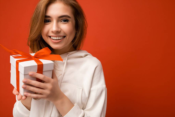 Beautiful happy young blonde woman isolated over colourful background wall wearing stylish casual clothes holding gift box and looking at camera - Photo, Image