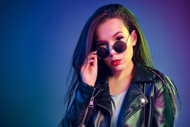 Asian girl in sunglasses and black leather jacket posing against dark blue background - 写真・画像