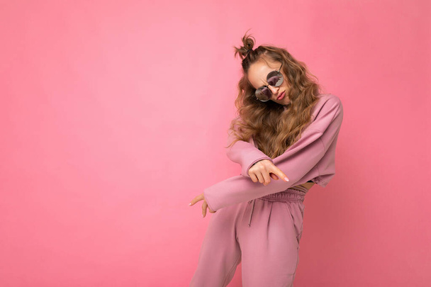 Prise de vue de belle jeune femme bouclée blonde sombre isolée sur un mur de fond rose portant des vêtements de sport rose décontracté et des lunettes de soleil élégantes regardant la caméra et dansant - Photo, image
