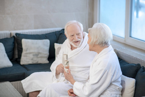 Mature couple in white robes sitting on the sofa and discussing something - Фото, изображение