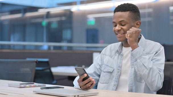 アフリカ人男性がオフィスでスマートフォンで成功を祝う  - 写真・画像