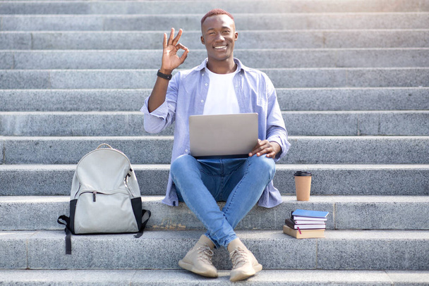 Online education. African American student with laptop, backpack and books showing okay gesture on stairs outdoors - Foto, Imagem