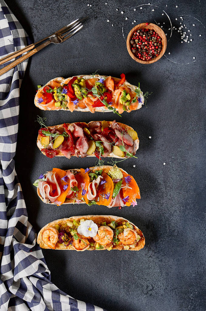 Italian Bruschetta antipasto - Grilled Bread topped with tomato, vegetables, cured meat, salted fish. Various bruschetta on dark textured table top view. Food flat lay, italian rustic dish concept - Foto, Imagem