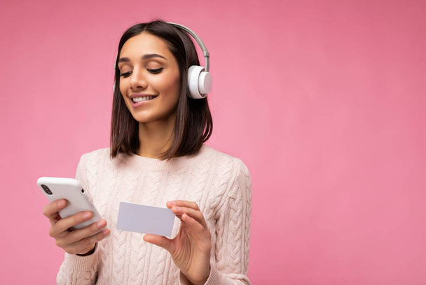 美しい肯定的な笑顔の写真ピンクのカジュアルなセーターを身に着けている若いブルネットの女性白いブルートゥースワイヤレスヘッドフォンを身に着けているピンクの背景の壁に隔離され、音楽を聴き、使用して - 写真・画像