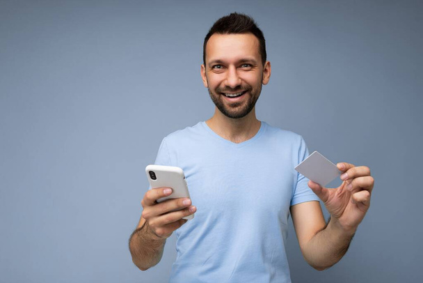 Usmívající se pohledný muž nosí každodenní oblečení izolované na pozadí zeď držení a používání telefonu a kreditní karty platby při pohledu do kamery - Fotografie, Obrázek