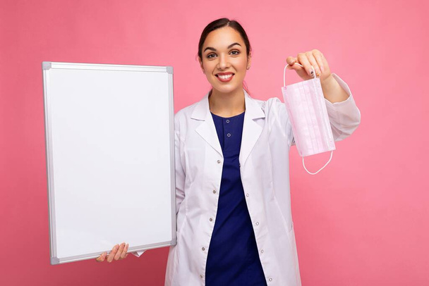 Vrouwelijke arts in een witte medische jas houdt blanco bord met kopieerruimte voor tekst en beschermende masker geïsoleerd op de achtergrond. Covid-concept - Foto, afbeelding