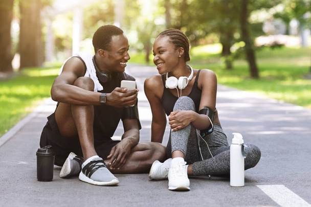 フィットネストレーニングの後にスマートフォンで休憩黒スポーツカップルの笑顔屋外 - 写真・画像