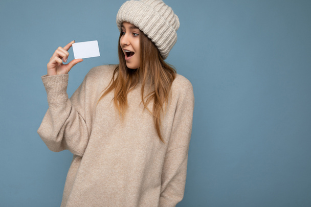 Красивая шоковая положительная улыбающаяся молодая темноволосая блондинка в бежевом свитере и вязаной бежевой шляпе на синем фоне, держащая и показывая кредитную карту глядя на пластиковый бесконтактный банк - Фото, изображение