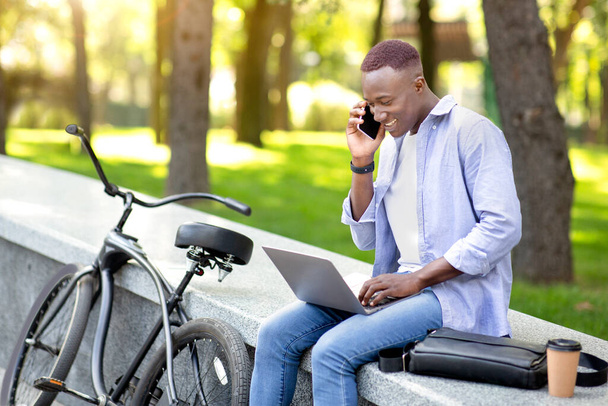 オンライン作業、教育、コミュニケーション。黒男と自転車で話している携帯電話や公園でラップトップを使用して、フリースペース - 写真・画像