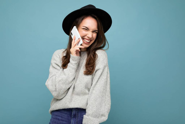 Attractive young smiling laughing woman wearing black hat and grey sweater holding smartphone looking at camera isolated on background - Photo, Image