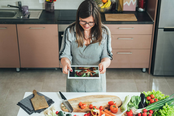 Piękna kobieta robi zdjęcia świeżych warzyw na bard cięcia przy użyciu tabletu cyfrowego. Uśmiechnięta gospodyni domowa krojąca świeże warzywa, trzymająca cyfrowy tablet i robiąca zdjęcia w kuchni - Zdjęcie, obraz