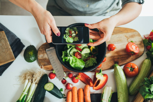 Asztali nézet egy nőről, aki friss salátát fényképez az asztalon, tele bio zöldségekkel. Felső fénykép háziasszony, hogy fotó ebéd friss színes zöldségek a konyhában otthon, vegán ételek, egészséges étkezési koncepció - Fotó, kép