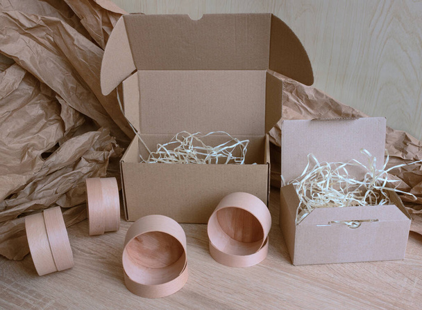 Cardboard boxes of different sizes and colors with natural filling, a large piece of wrapping paper, four small round boxes on a wooden background. Eco-friendly packaging for cosmetics and beauty salons. - Photo, Image