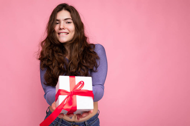 Shot of attractive positive smiling young brunette woman isolated over colourful background wall wearing everyday trendy outfit holding gift box and looking at camera - Photo, Image