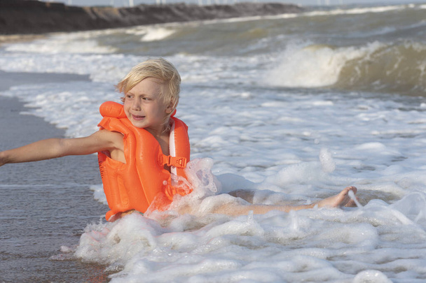 Vaaleahiuksinen poika puhallettavassa uimaliivissä istuu meren rannalla vedessä - Valokuva, kuva