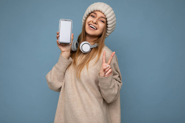 Fotografie krásné šťastný úsměv mladá tmavá blondýna žena na sobě béžový stylový svetr a pletená béžová zimní klobouk izolované na modrém pozadí na sobě bílé bluetooth bezdrátové sluchátka a zobrazení - Fotografie, Obrázek