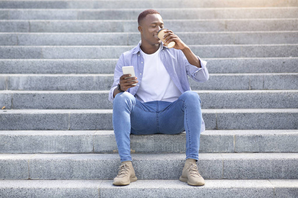 外の階段に座っている間にテイクアウトコーヒーを飲み、携帯電話を使用してハンサムな黒人男性 - 写真・画像