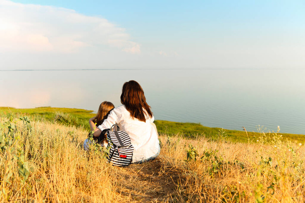 Vista posteriore di donna irriconoscibile abbracciare ragazza mentre seduto su erba secca e ammirare pittoresco paesaggio marino durante il fine settimana estivo - Foto, immagini