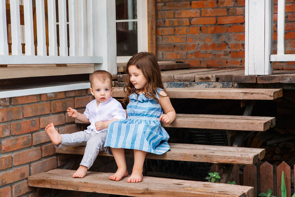Lindo hermano pequeño y hermana sentado en la escalera de madera cerca del porche de la casa de campo y relajarse en la zona rural durante las vacaciones de verano - Foto, Imagen