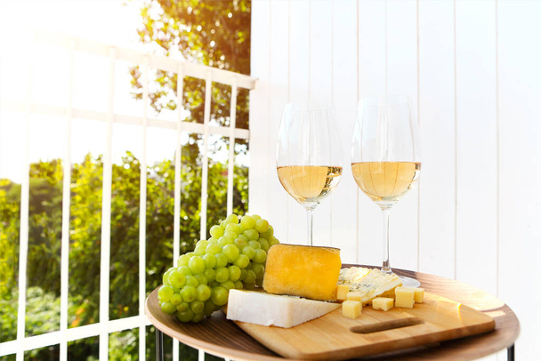 Güneşli bir günde terasta olgun üzüm ve peynirle kesme tahtasının yanındaki masaya şarap kadehleri konur. - Fotoğraf, Görsel