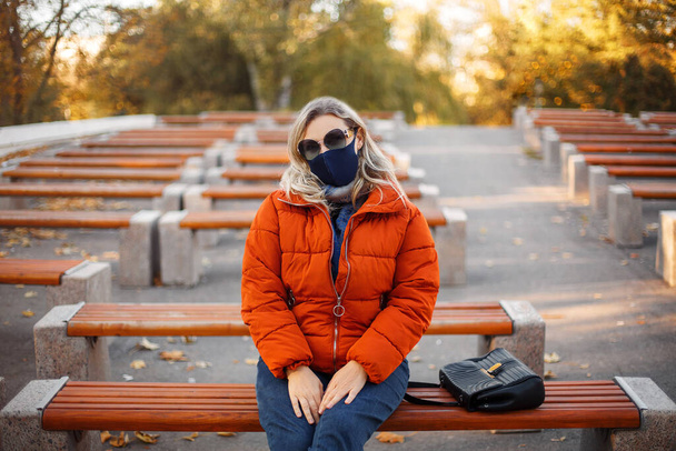 Femme blonde en vêtements de dessus élégants et masque en tissu regardant la caméra tout en piquant sur le banc dans le parc pendant la pandémie - Photo, image
