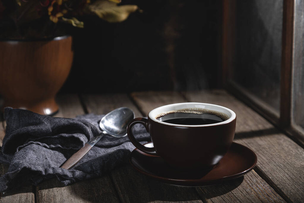 Cup of hot coffee next to a window on a rustic wooden table - 写真・画像