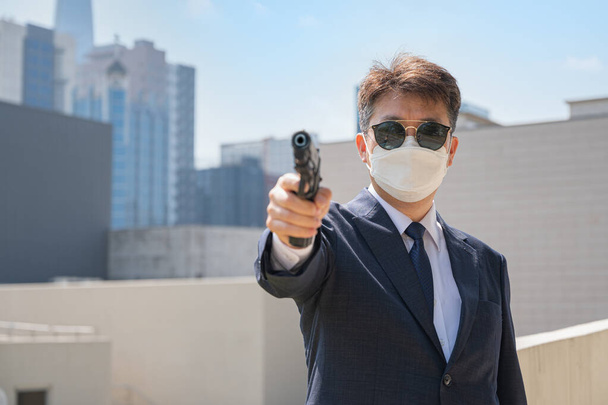 Ασιάτης άνδρας που κρατάει πιστόλι φορώντας γυαλιά ηλίου και μάσκα στο βάθος της πόλης. - Φωτογραφία, εικόνα
