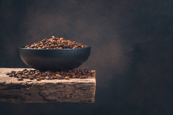 Black ceramic plate with coffee beans on a wooden board on a brown background. Side view, copy space. - Photo, Image